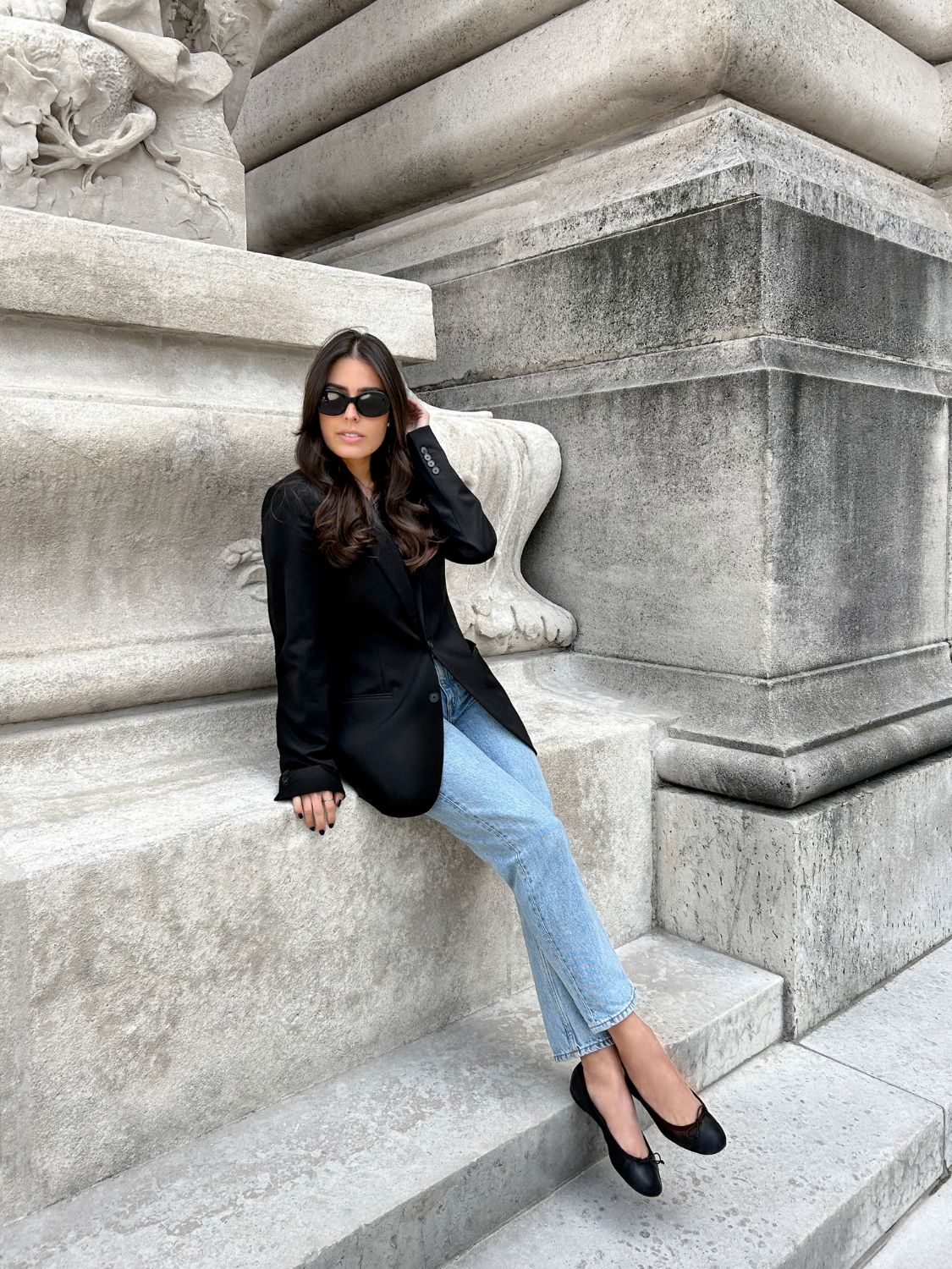 Lissa Barquero in an oversized black blazer and ballet flats, seated on stone steps